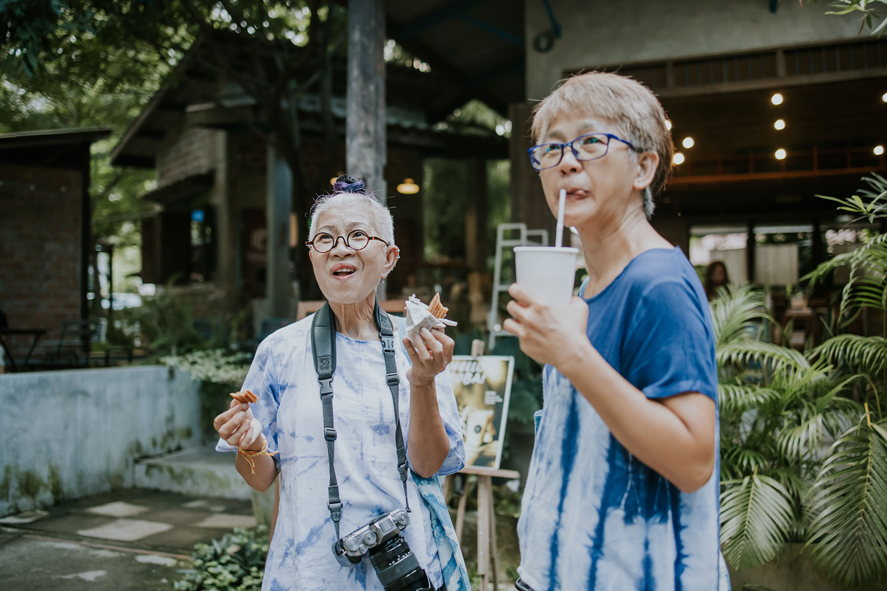 Two happy older ladies standing outside of a local restaurant or cafe  in the Chiangmai province. One is eating a snack, the other is drinking a drink from a single-use cup.
