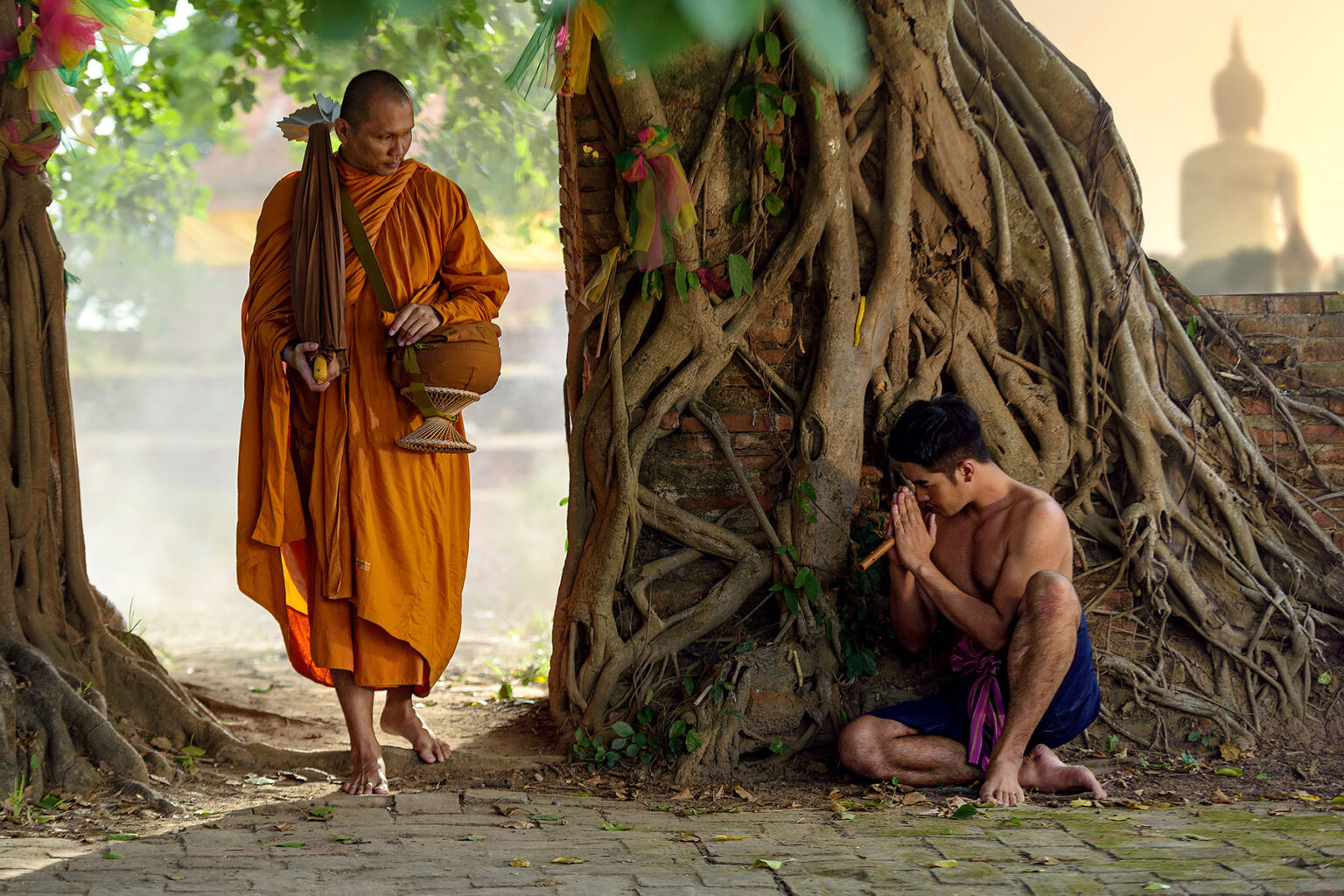 Man sitting on the ground near a tree, and greets a passing monk with a traditonal Thai wai. A large statue of Buddha looms in the background.