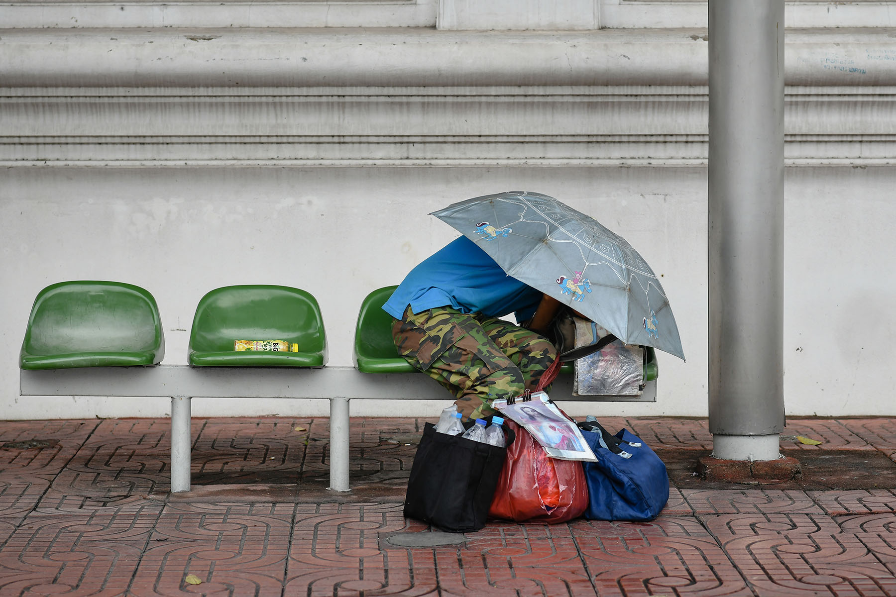 A houseless person is hiding underneath an umbrella while sitting near a bus stop in Bangkok.