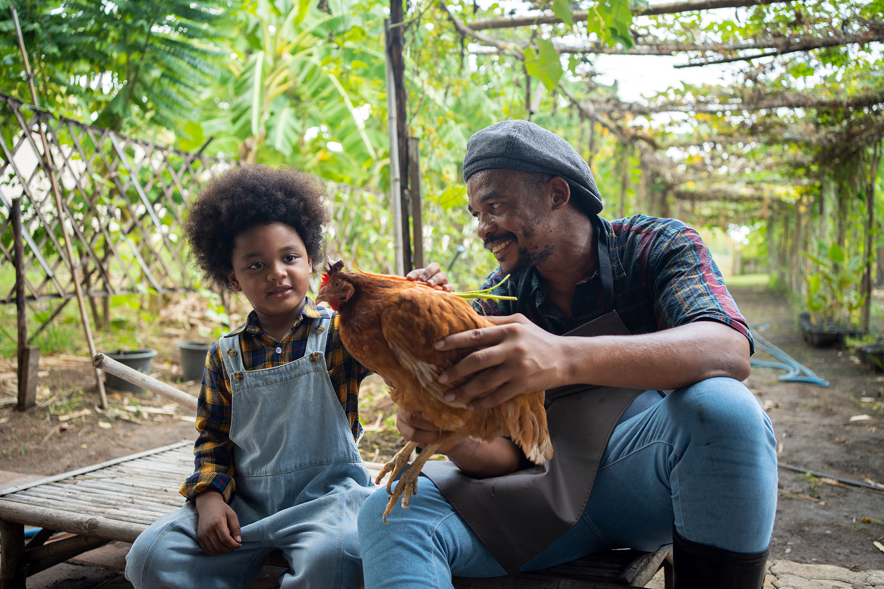 Self-employed farmer is holding a chicken while smiling and looking at a child beside them, who is petting the animal.