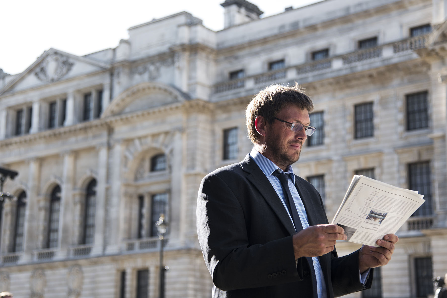 Man with glasses reading a newspaper outside a large stately-looking building.