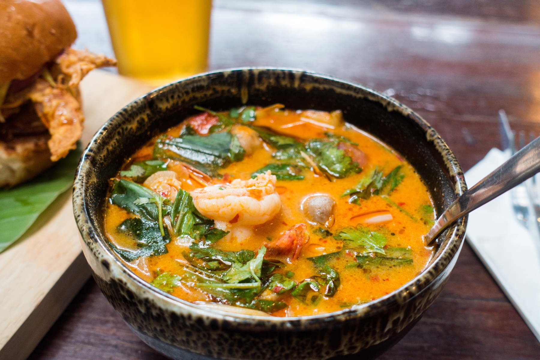 A bowl of tom yum goong with added crab, herbs, alongside a burger and a beer