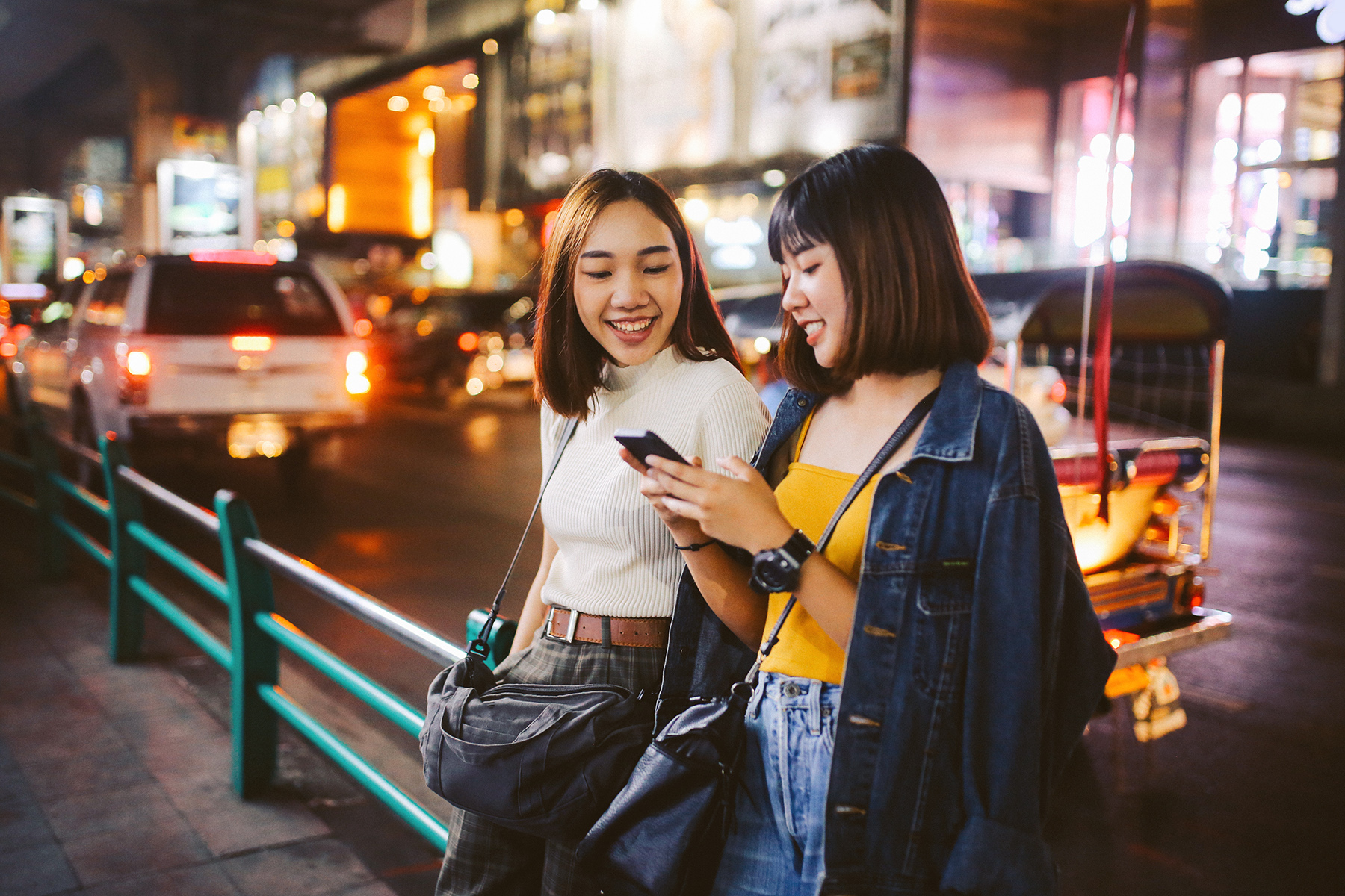 Two young women leaning against a sidewalk railing, smiling and texting on their phone

