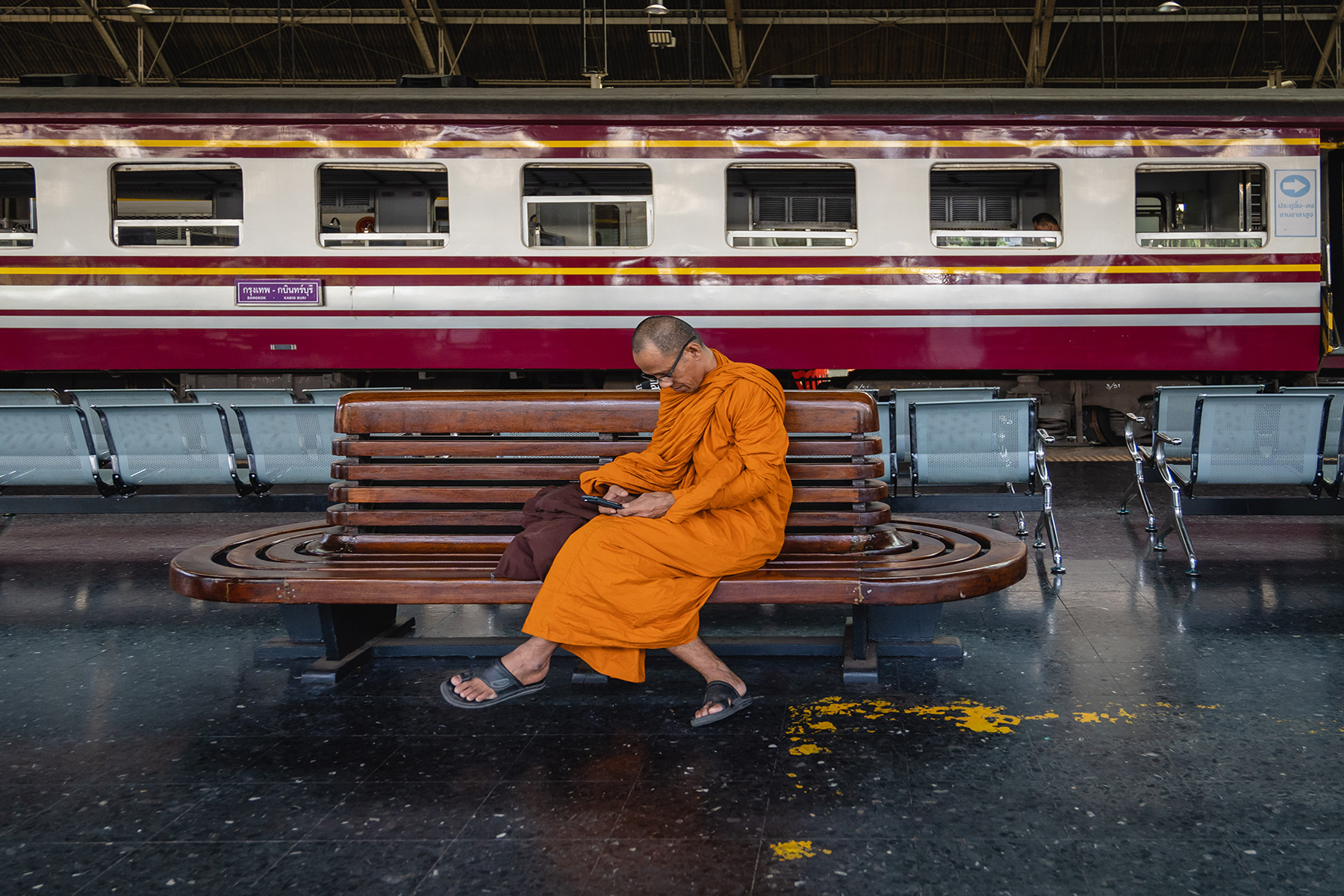 A Thai monk in orange robes sits at Hua Lamphong Station the train station and looks at his mobile phone
