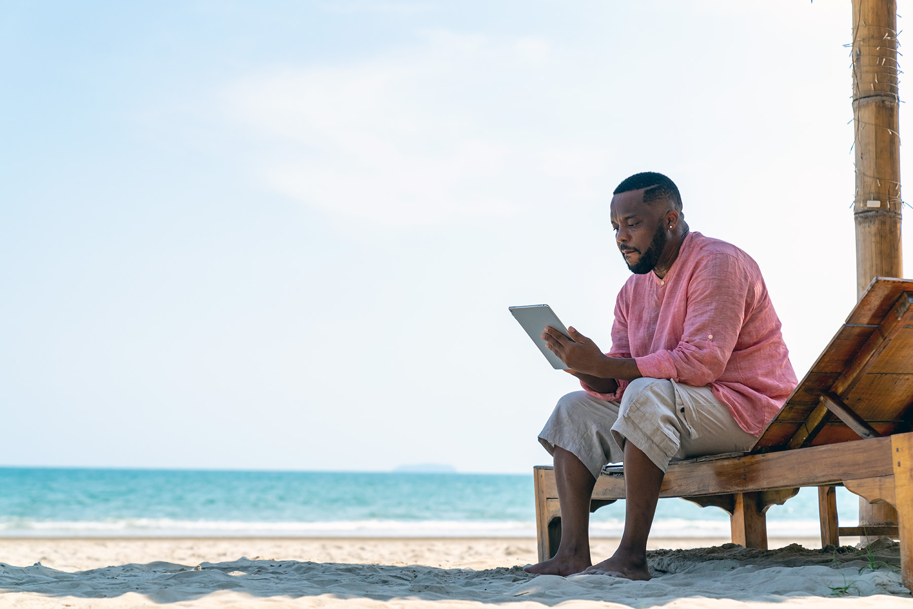 Man looking pensively at his tablet while sitting on a pullout beach chair on the beach in Thailand.