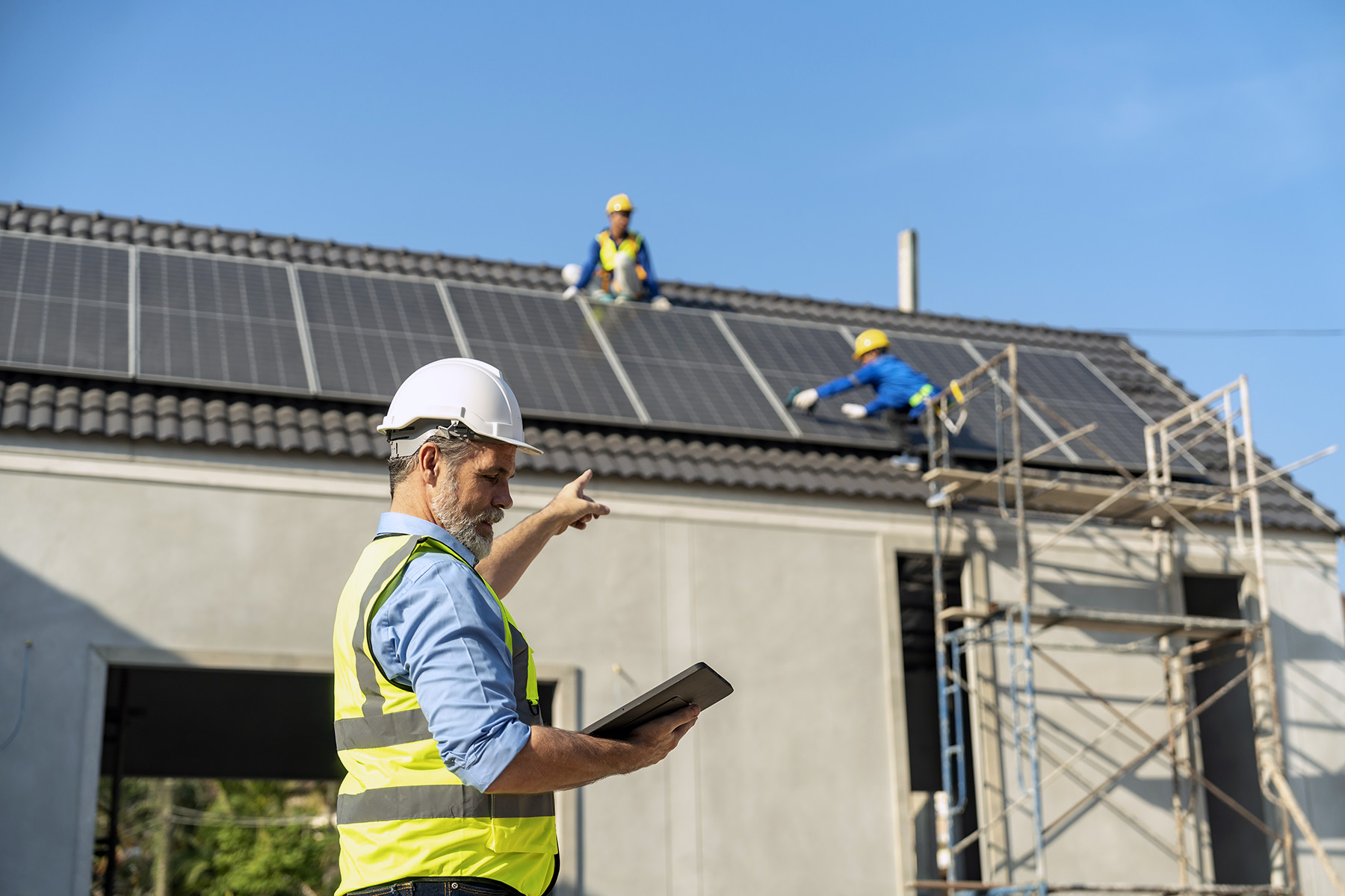 Man in hi vis vest and white safety hard hat holding a tablet while he is directing two workers working on installing solar panels on the roof of a house.
