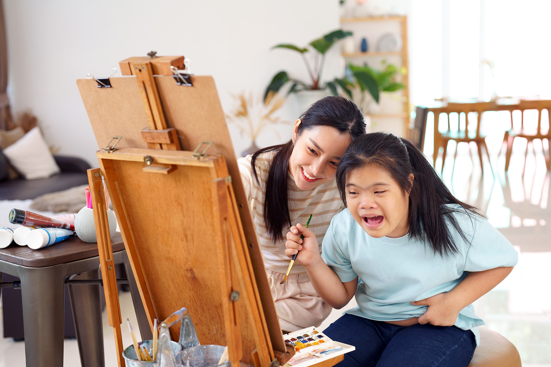 Happy smiling mother and daughter painting using an easel. The child has down syndrome and is holding the brush wrong. Still, they're having fun.