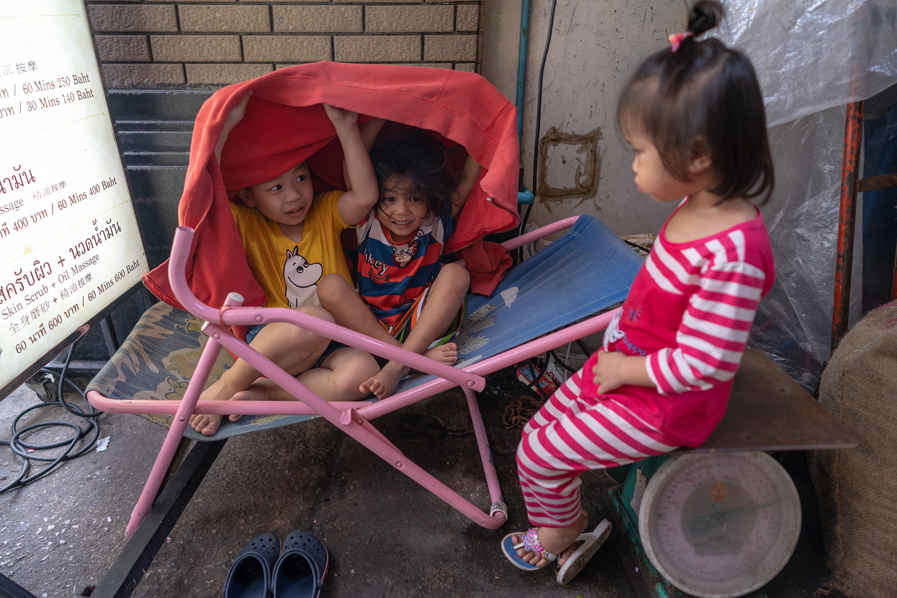 Two girls playing hide and seek to entertain a third, younger girl. In typical younger sister fashion, she is not and likely more angry she isn't allowed in the wheelbarrow.