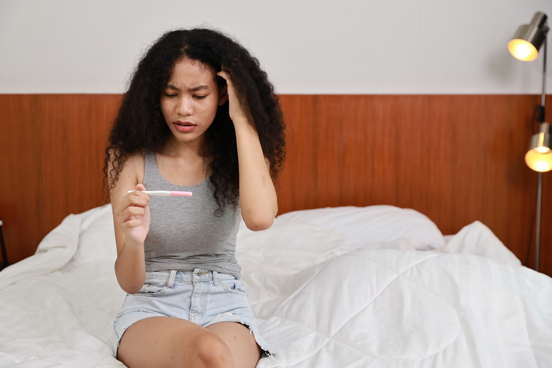 Pensive looking young woman sitting on a bed, while scratching her head and looking at a pregnancy test.