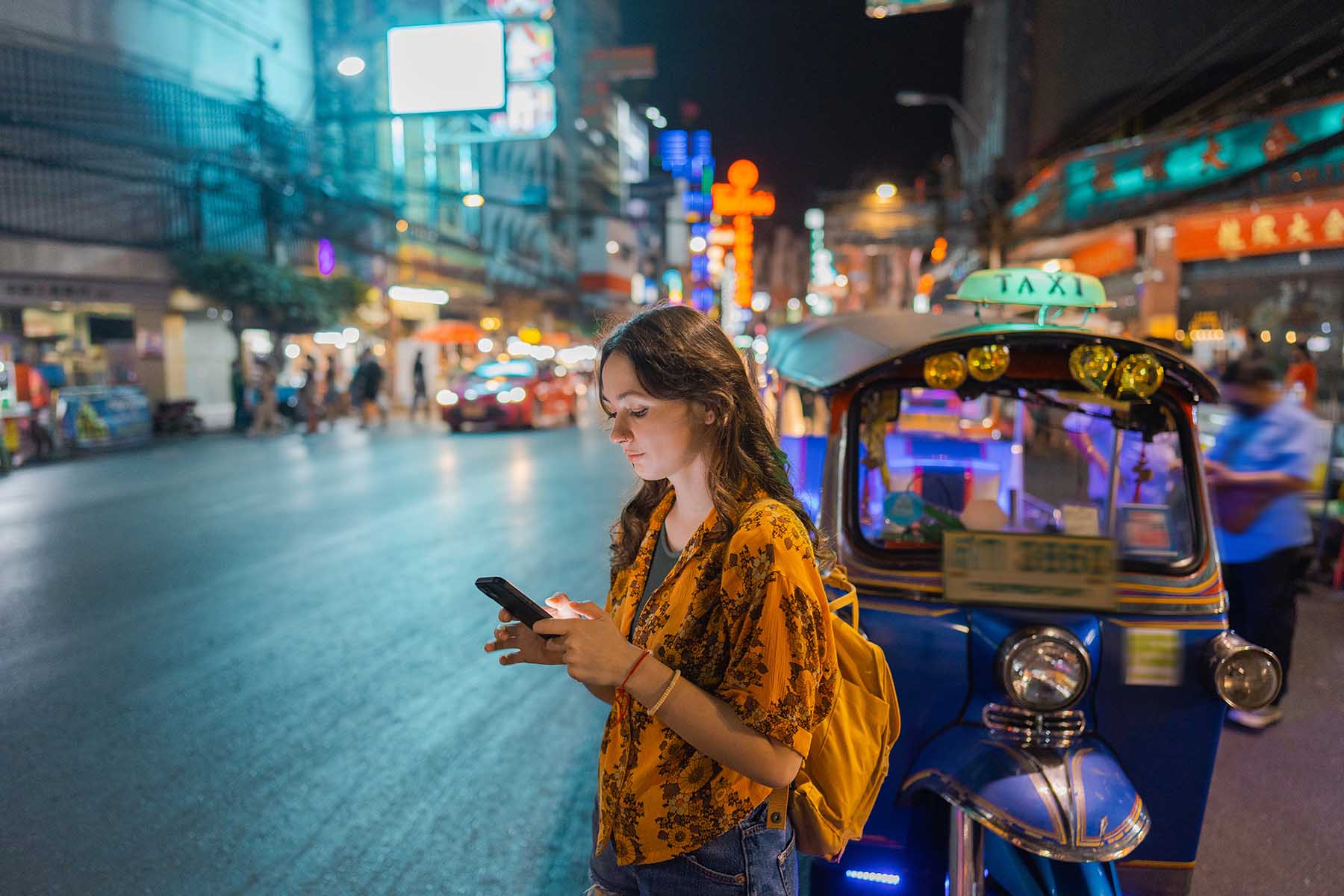 Woman looks at her mobile phone while standing in front of a tuk tuk at night on a street in Bangkok, Thailand.
