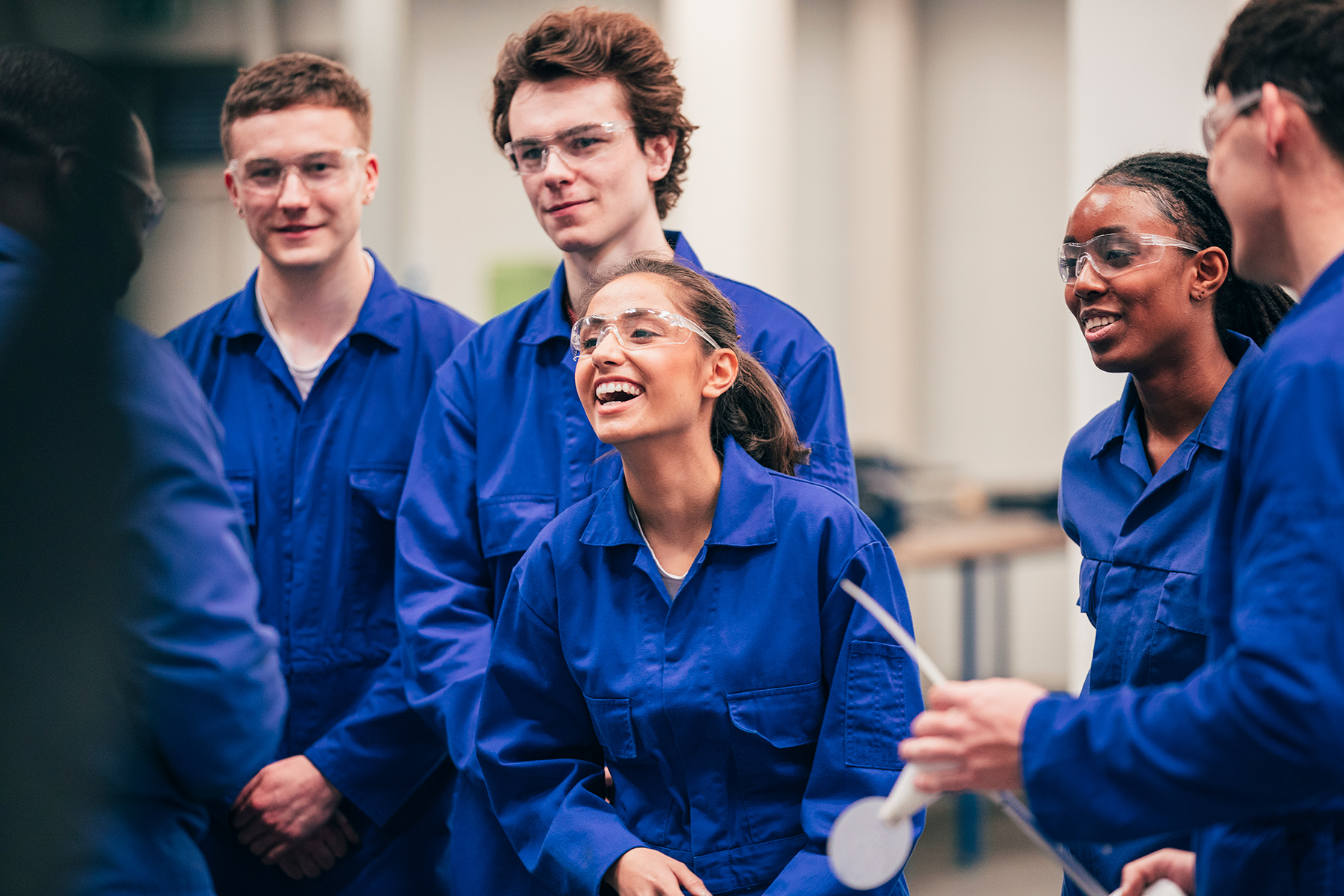 A group of young apprentices in blue boiler suits laughing. Some are wearing goggles.