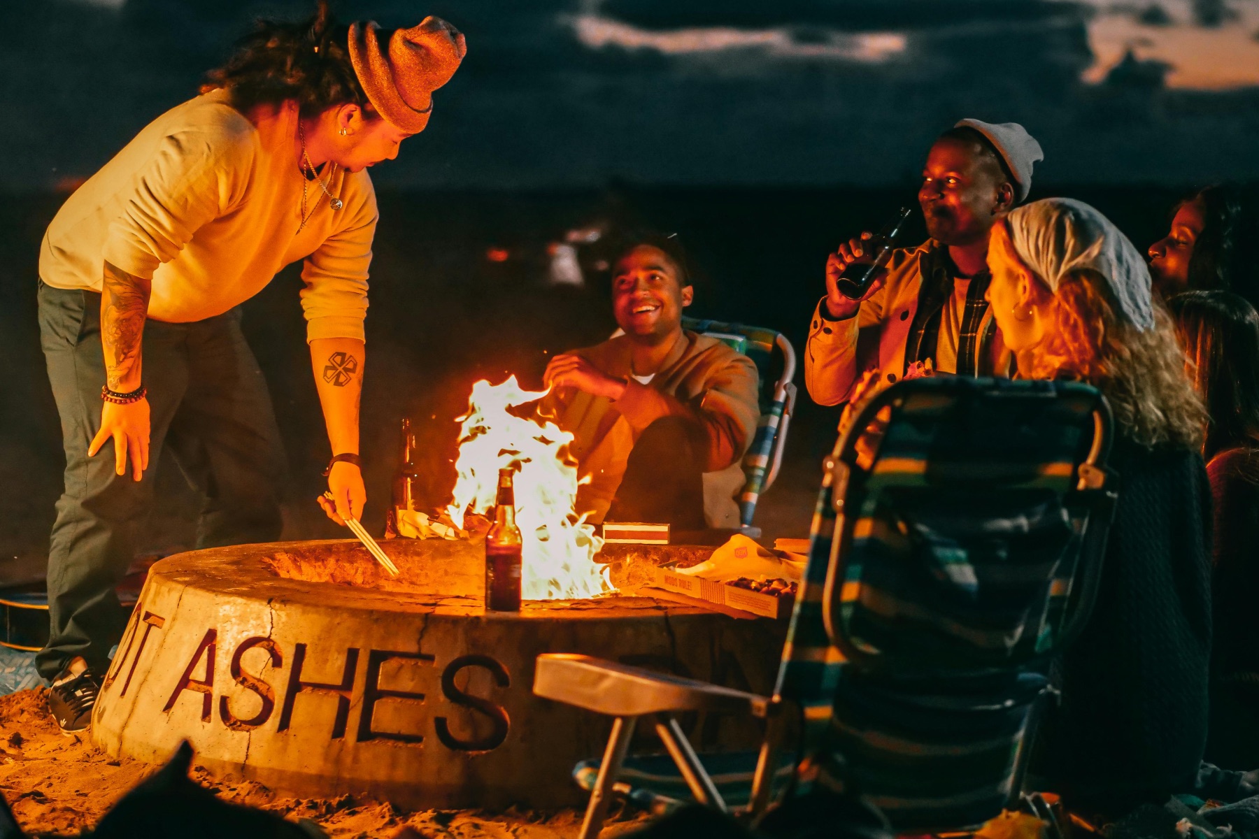 A group of friends in their 30s gather around a bonfire on a beach, smiling and drinking beer