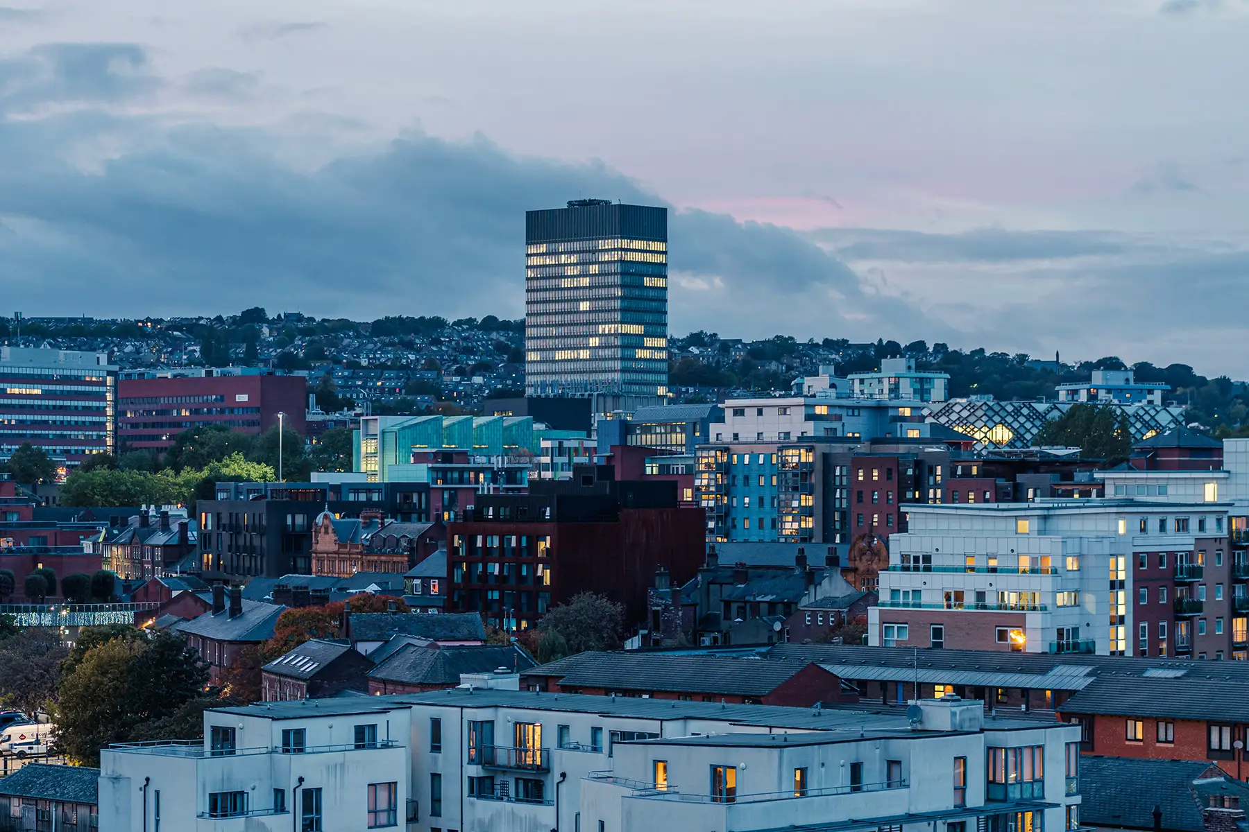 Sheffield cityscape centered on a tall building at dusk with lots of lights on in different buildings.