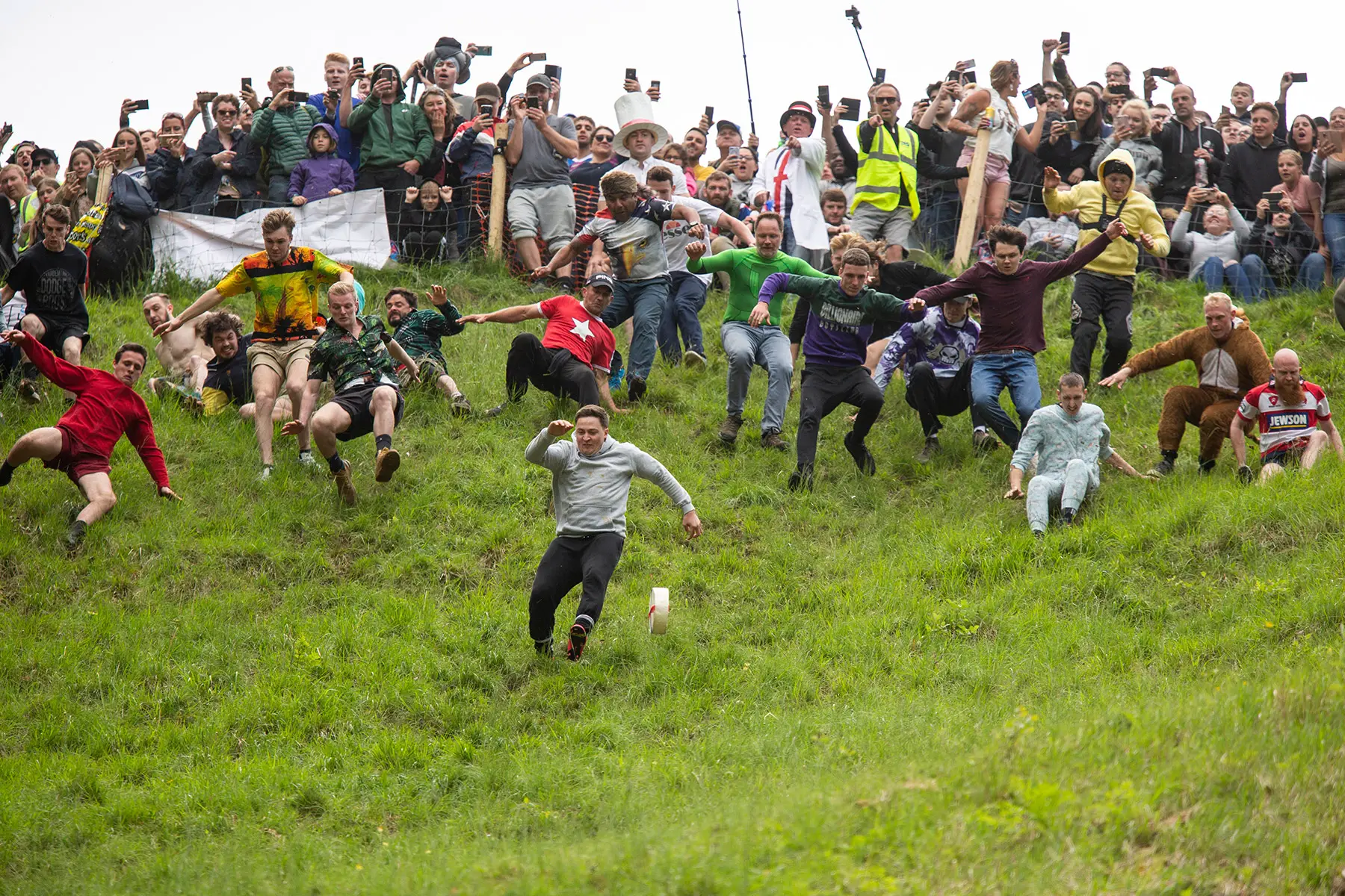 A crowd of men chase a wheel of cheese down a steep hill