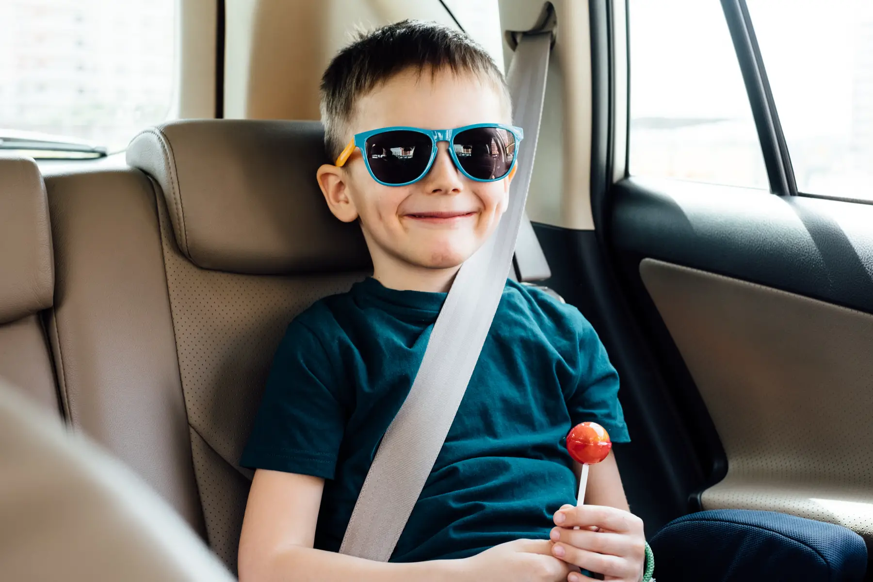 a child with a lollipop and sunglasses sitting in the back seat of a car wearing a seatbelt