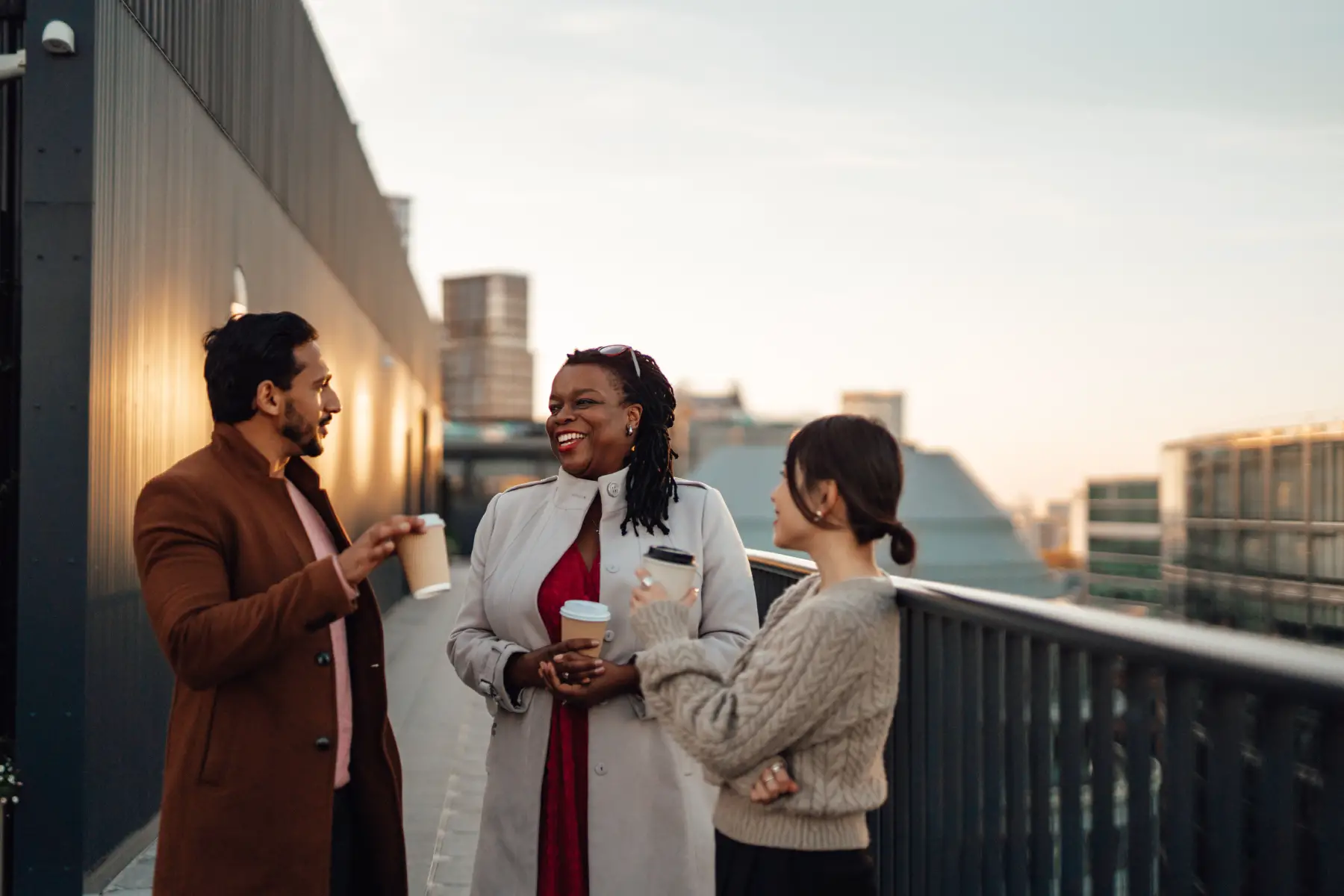 Three colleagues laugh on rooftop while drinking coffee