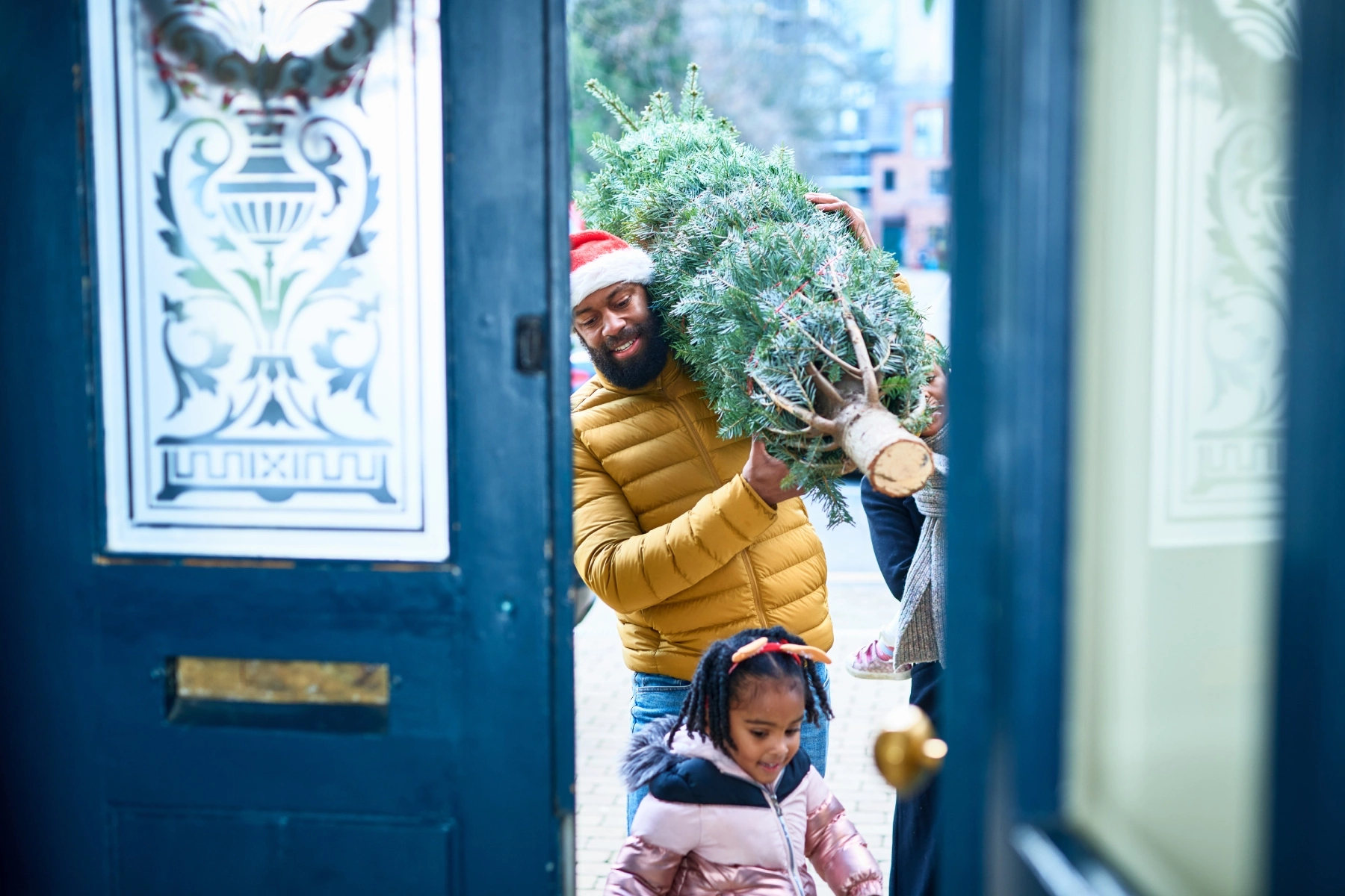 Father carrying Christmas tree and daughter enter their home in London