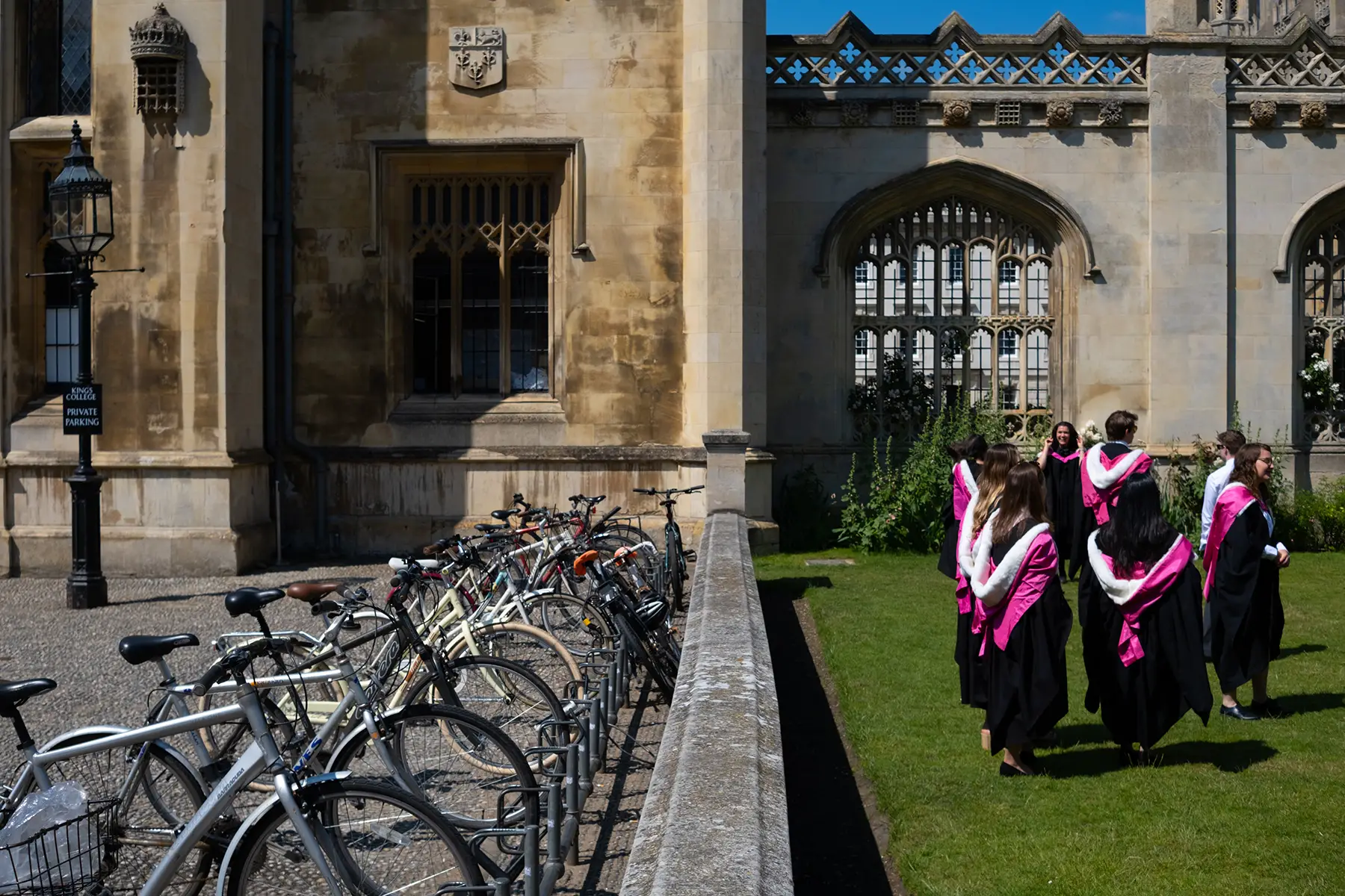 University of Cambridge students in graduation robes standing on grass outside a university building next to a row of parked bicycles.