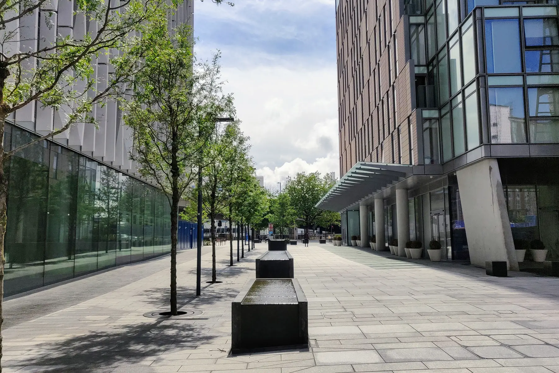 A paved pedestrian area with tall glass buildings either side and a fountain in the middle.