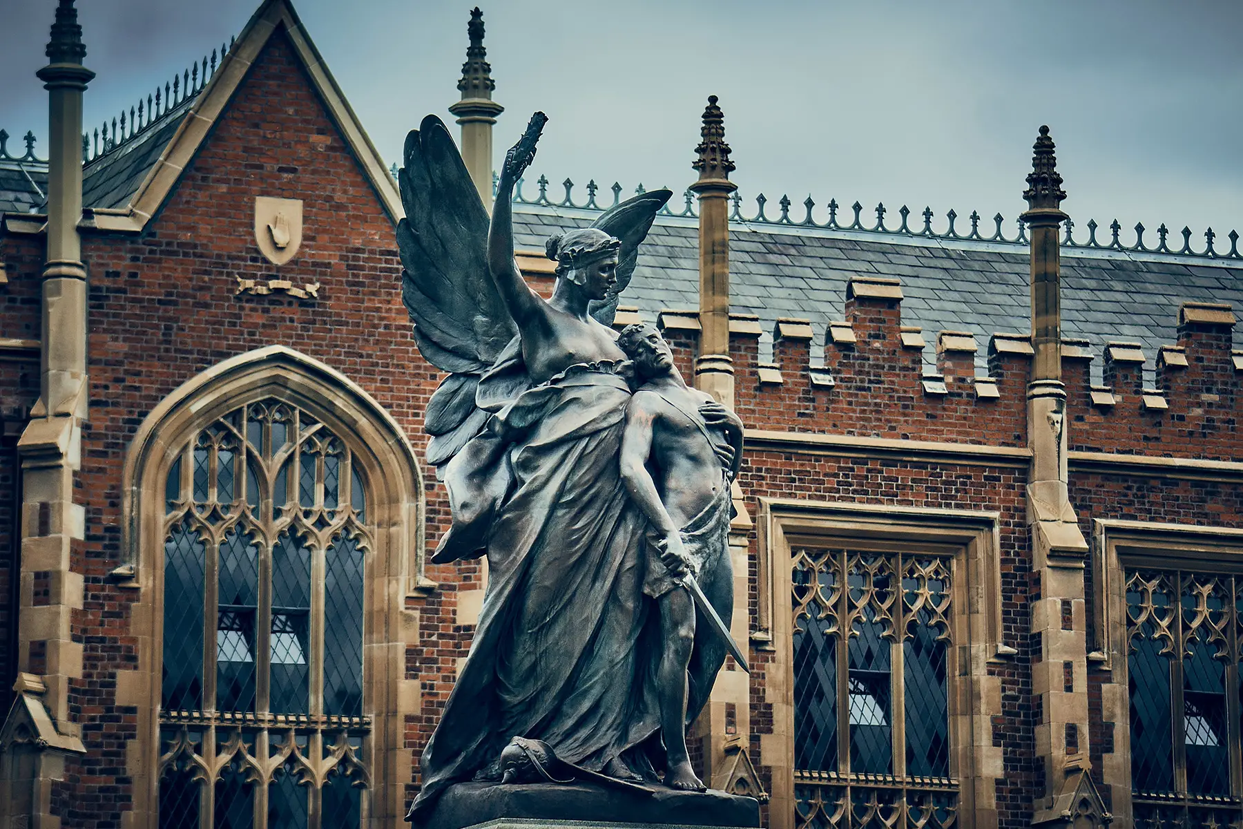 Statue of an angel behind a man with a sword. The statue is in front of a gothic-style university building.