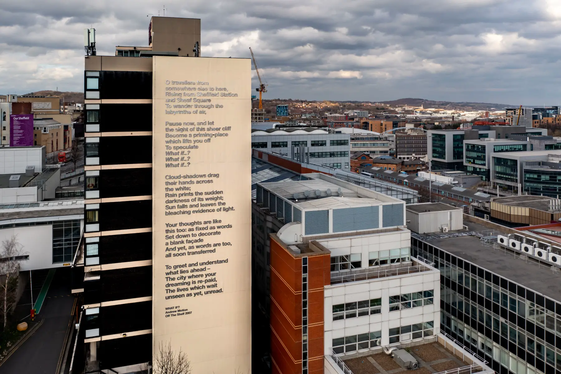 A modern university building at Sheffield Hallam university, with Sheffield skyline as background. There is a poem on the side of the building: What If? by Andrew Motion.