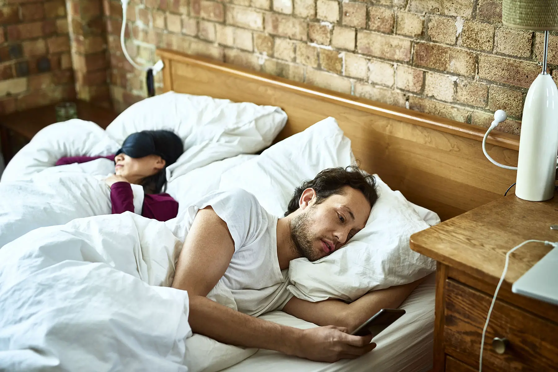 Man looking at his phone while his wife is sleeping next to him in the bed.