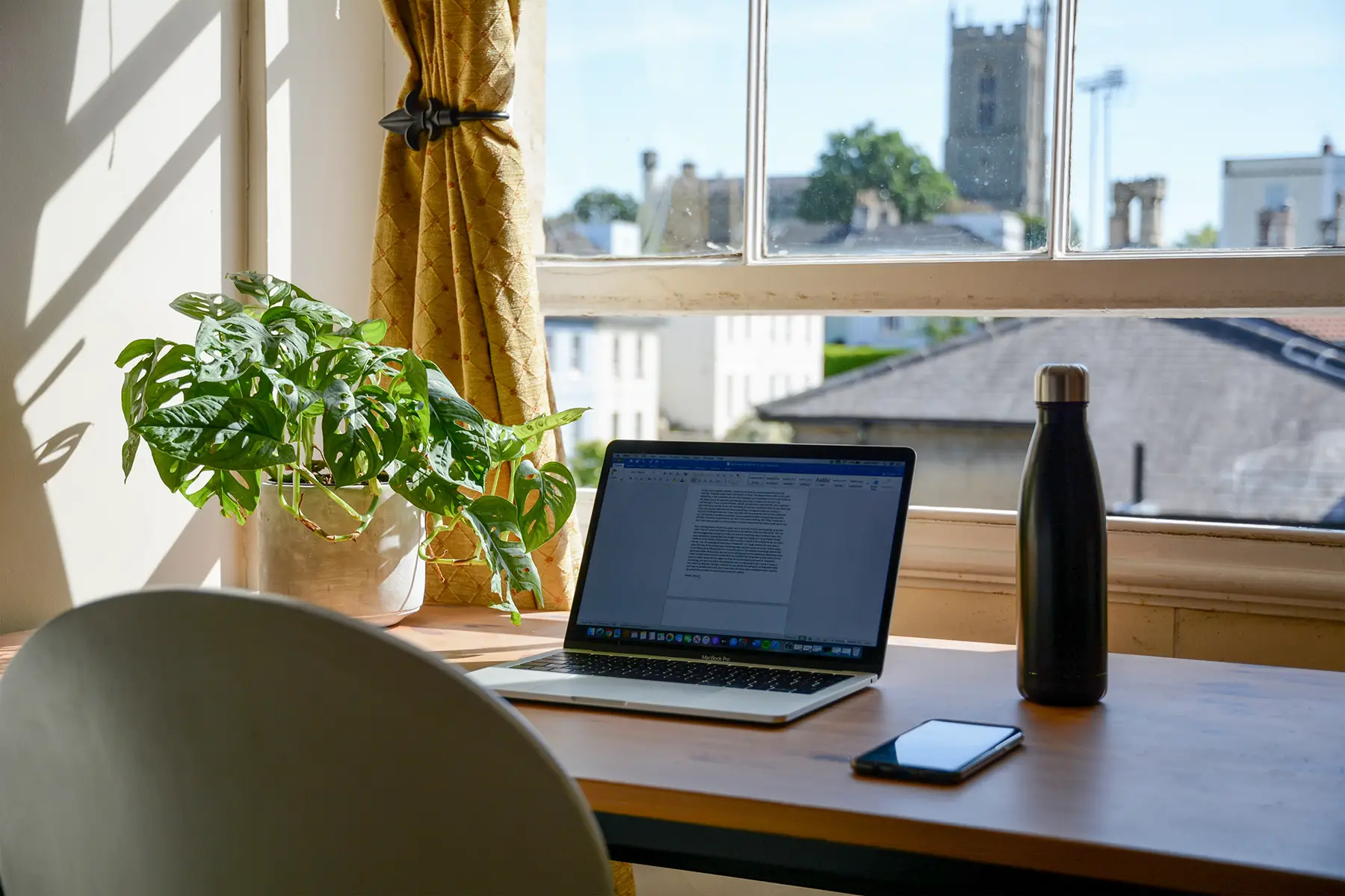 A desk with a laptop, bottle of water, and smartphone on it. The desk is by an open window showing a view of Bristol (old buildings including cathedral)