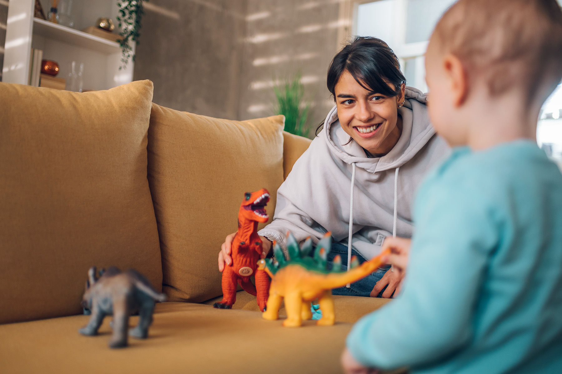 Au pair and toddler child playing with dinosaurs toys on a couch at home.