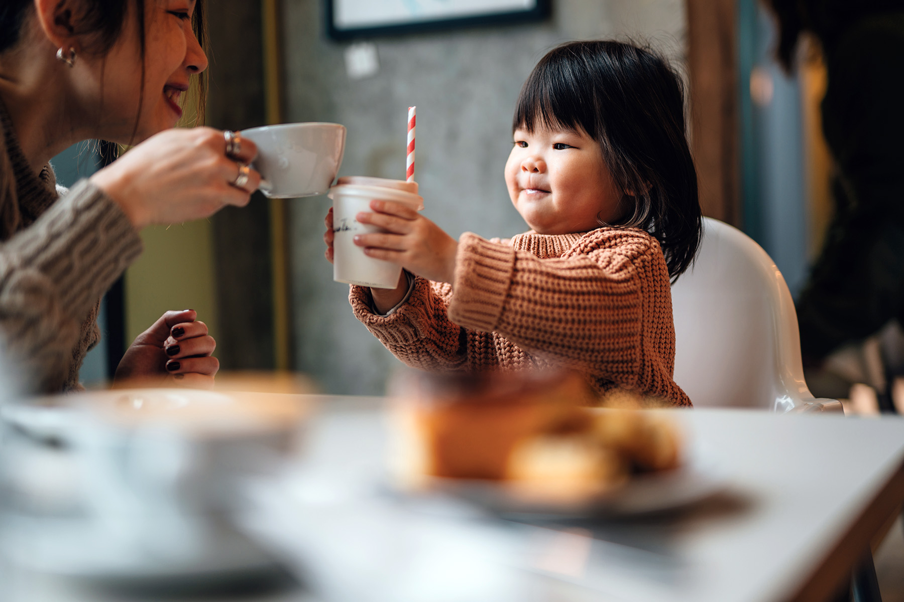 Toddler daughter is cheersing her mother's cup of coffee at a cafe.