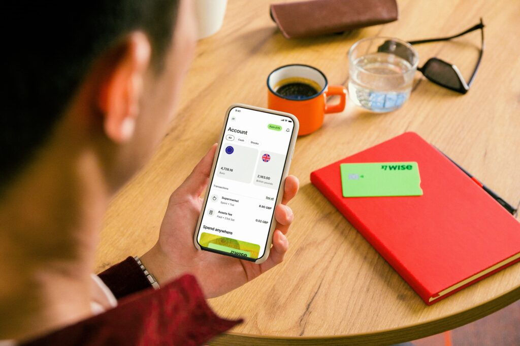 A man sits at a table looking at his Wise Account on his smartphone. Behind him on the table lays a Wise Card and other gadgets.