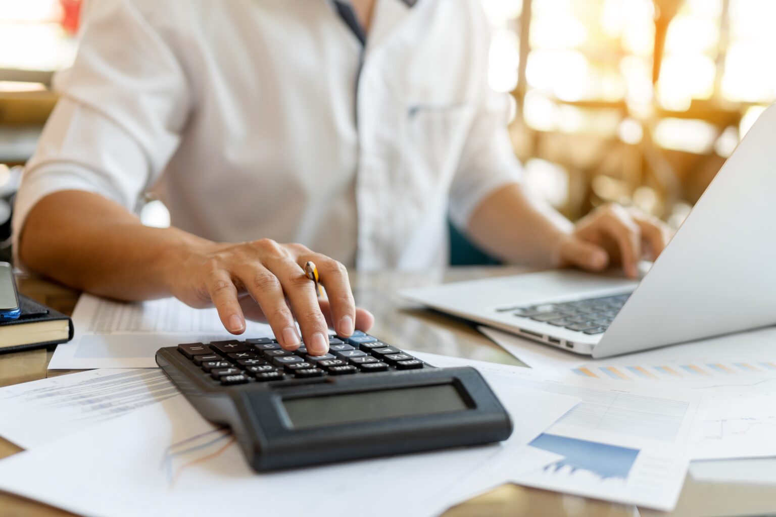 Man working on a laptop covered in paper bills and tipping numbers into a calculator next to it.