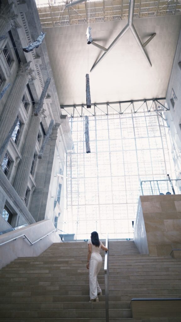 A woman seen from behind, dressed in white, walks up a grand stone staircase inside a bright, modern atrium. The space features massive classical columns, a soaring glass wall, and contemporary art installations suspended from the high ceiling.