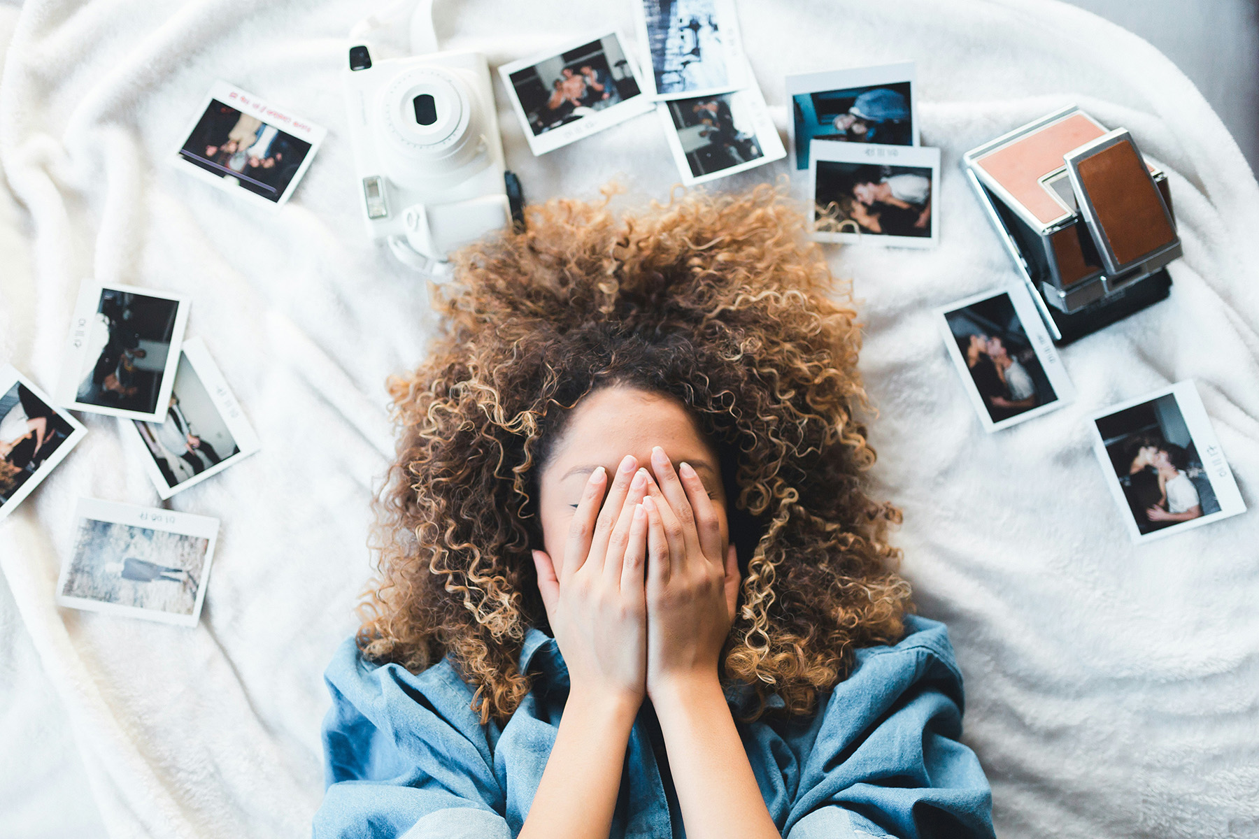 Girl lies on bed, coves her face with hands, photos scattered around