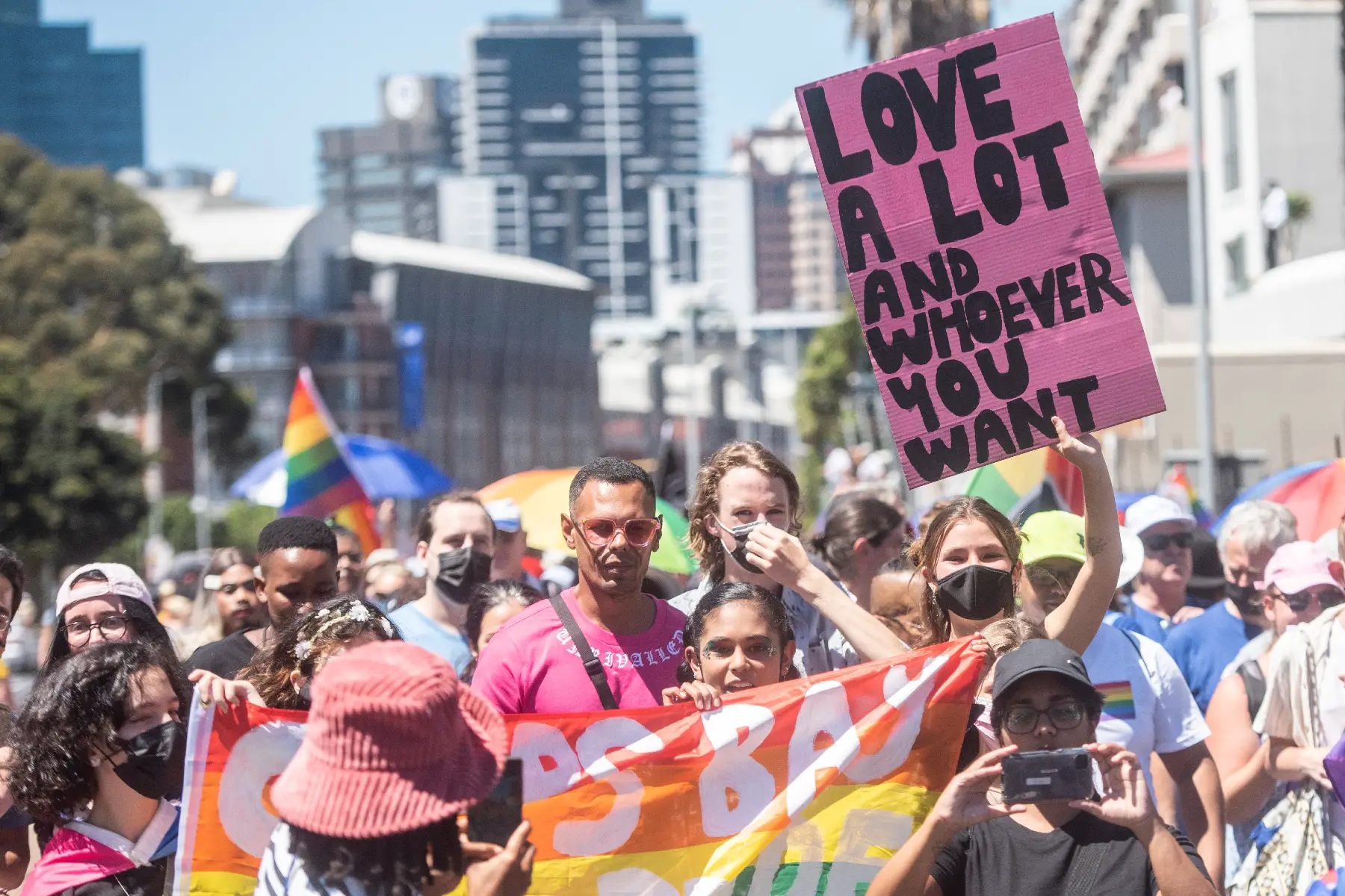 People marching behind a rainbow flag at Cape Town Pride 2022. A woman holds a pink sign saying 'Love a lot and whoever you want'