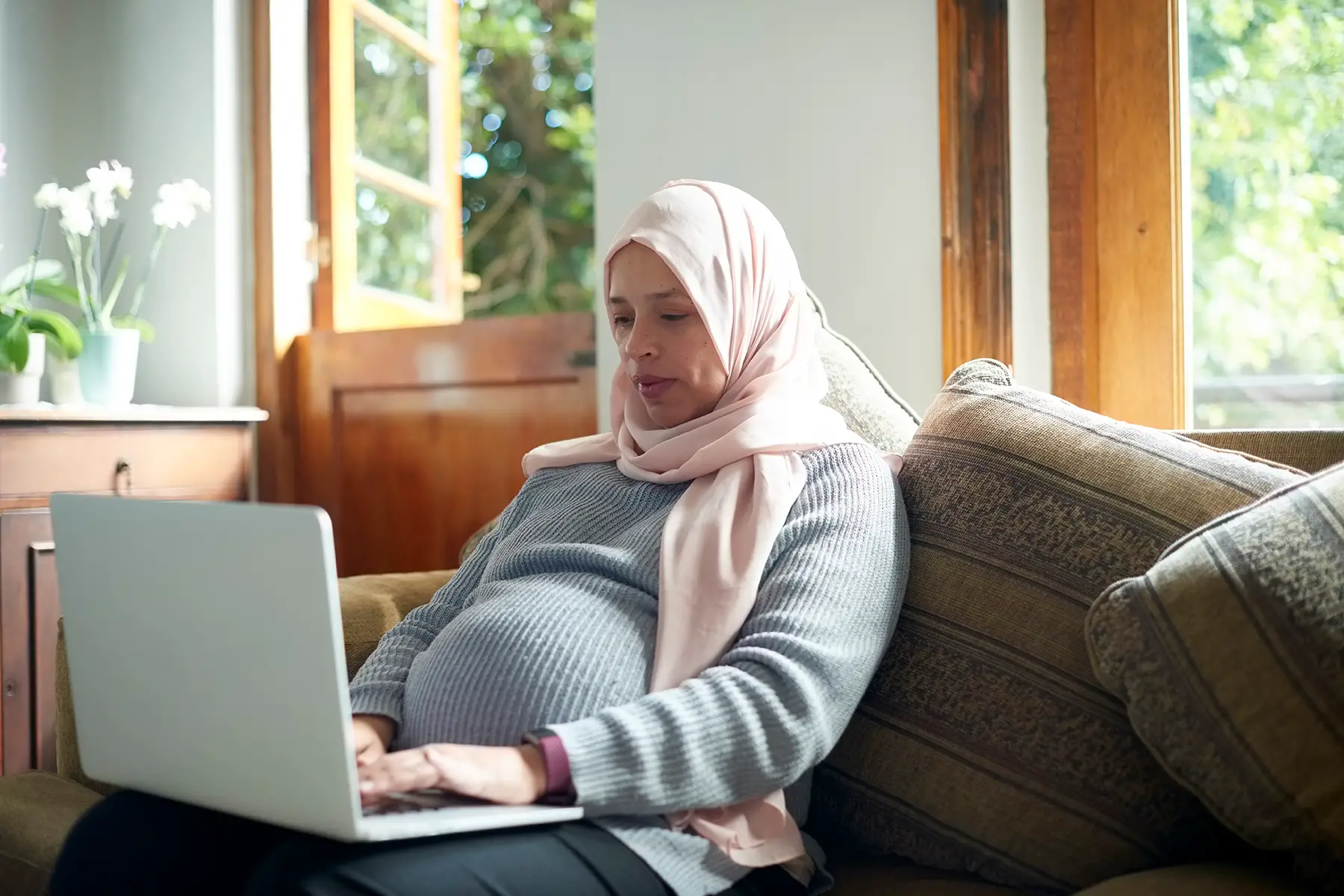 Muslim, pregnant woman with head scarve, sitting on couch using laptop to apply for her permanent residence in South Africa