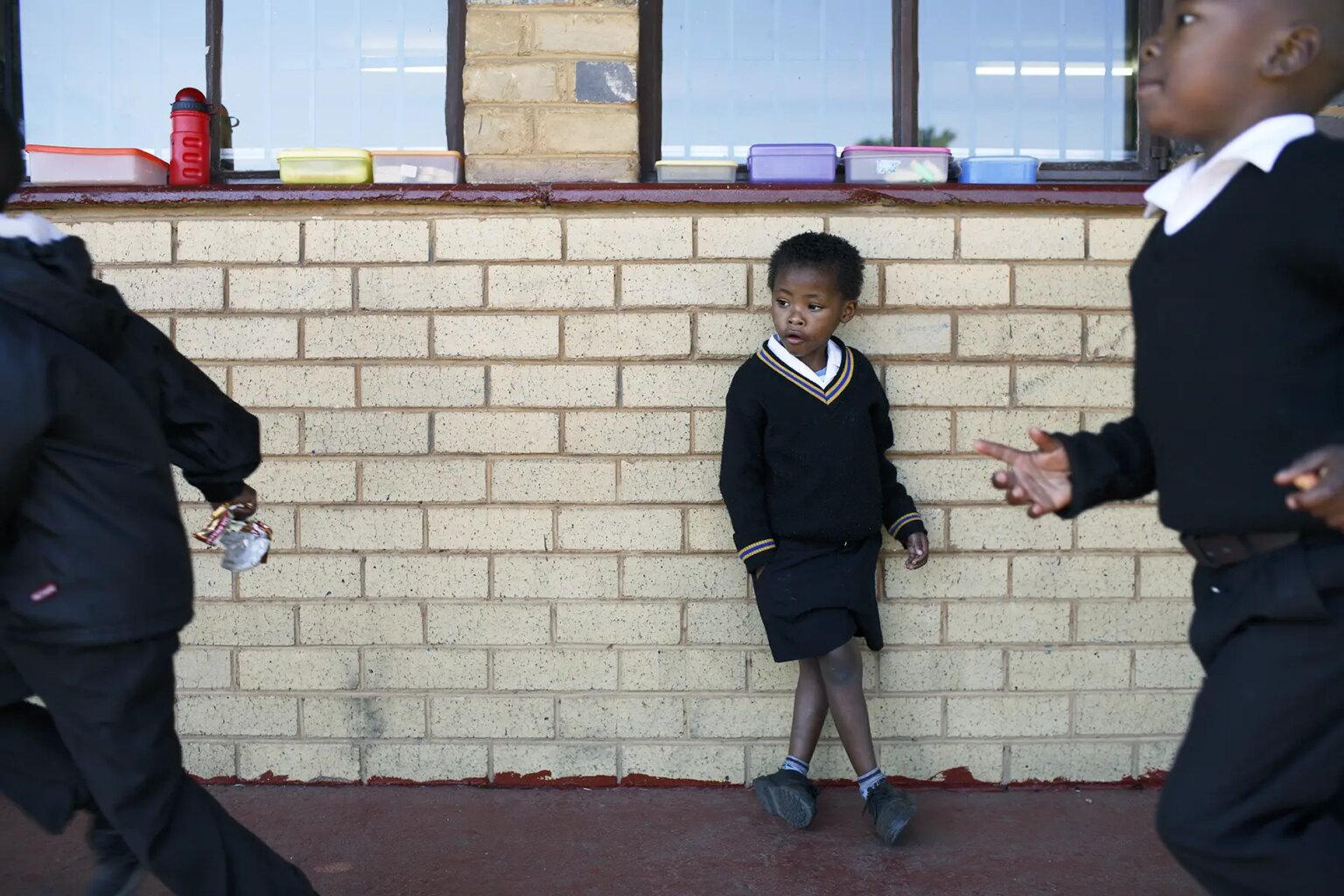 Young child in school uniform leans against a brick wall, other children run past