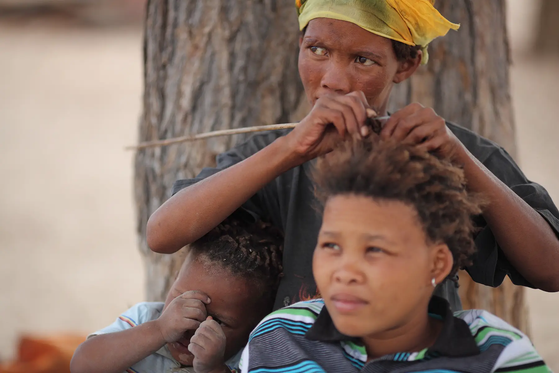 A San Bushwomen from the Khomani San community styles a young girls hair in the Southern Kalahari Desert. A young child sits next to them.