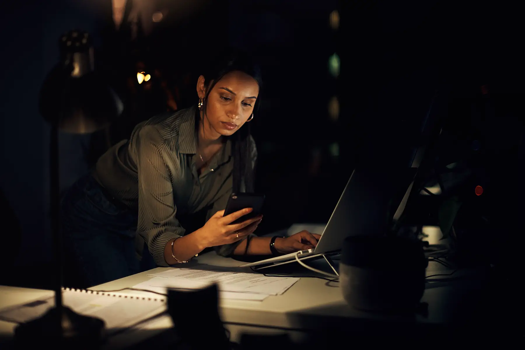 Woman working late, only lit by the desk lamp and the light from her laptop and phone, she looks irritated.