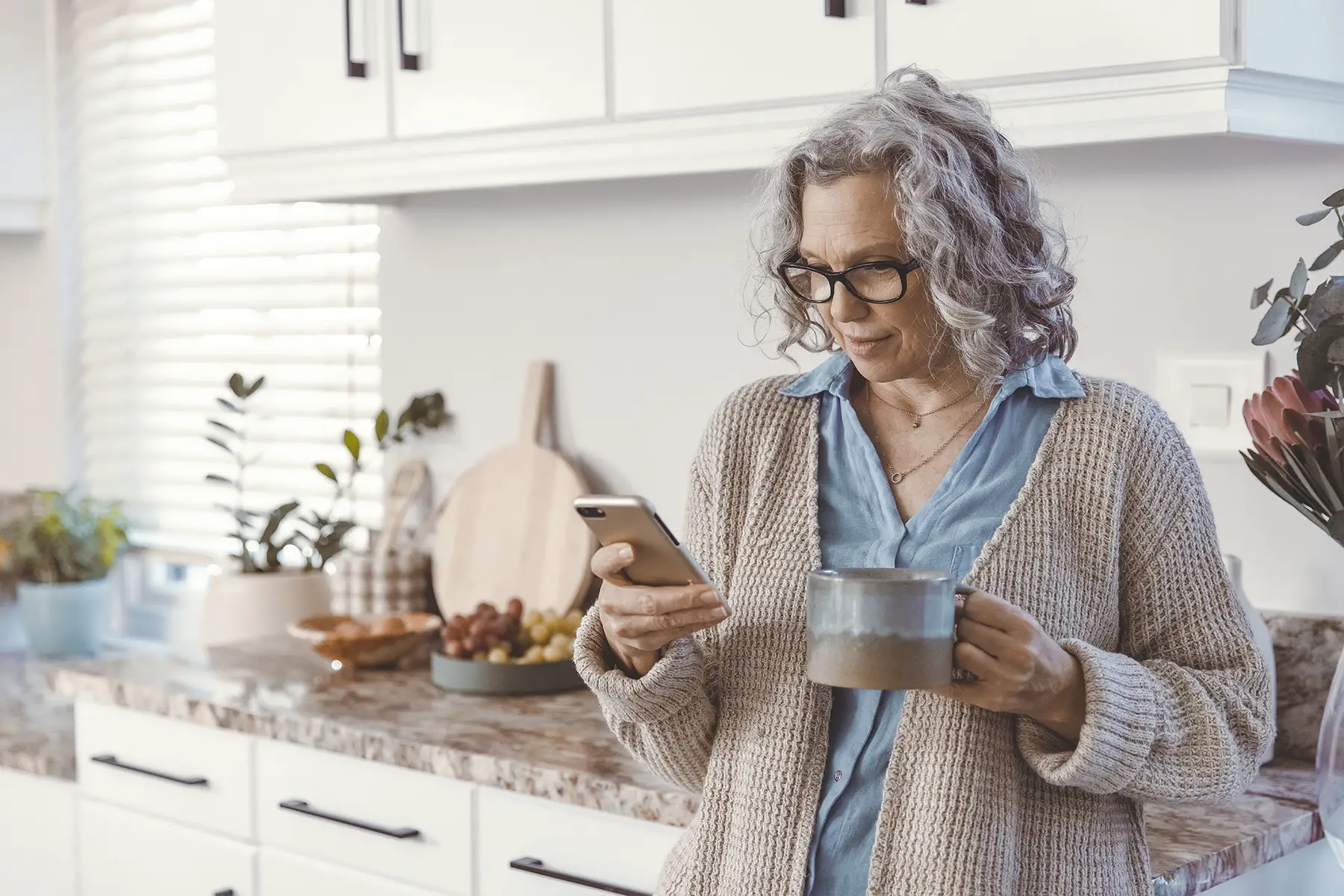 A woman looks at her phone, with a cup in her hand, while standing in her bright kitchen