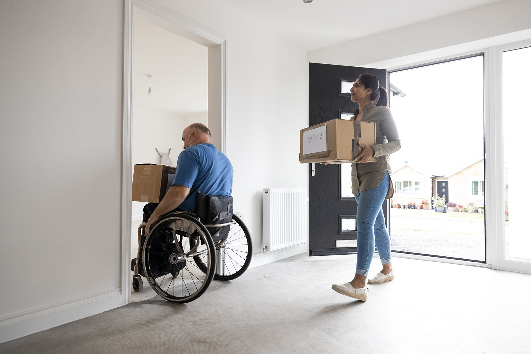 A man who uses a wheelchair goes into an empty, new house carrying a moving box, a woman walks with another box behind him